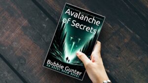 Hand holding a book titled 'Avalanche of Secrets' by Bubbie Gunter on a dark wood background