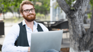 Confident bearded man in glasses and a casual vest working on a laptop outdoors, with a pleasant smile, suggesting productivity and enjoyment in a relaxed environment
